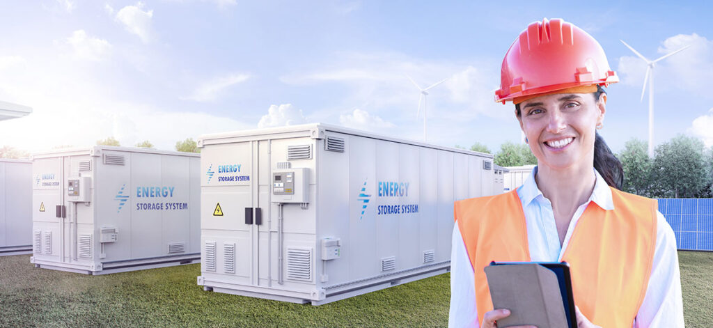 A female engineer or electrical technician wearing an orange vest and hard hat stands holding a notepad and smiling while working outdoors